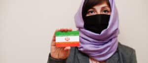 ortrait of young muslim woman wearing formal wear, protect face mask and hijab head scarf, hold Iran flag card.