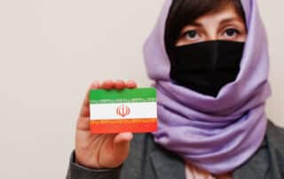 ortrait of young muslim woman wearing formal wear, protect face mask and hijab head scarf, hold Iran flag card.