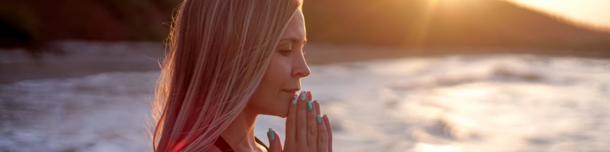 A female prays on a beach as the sun sets behind her.