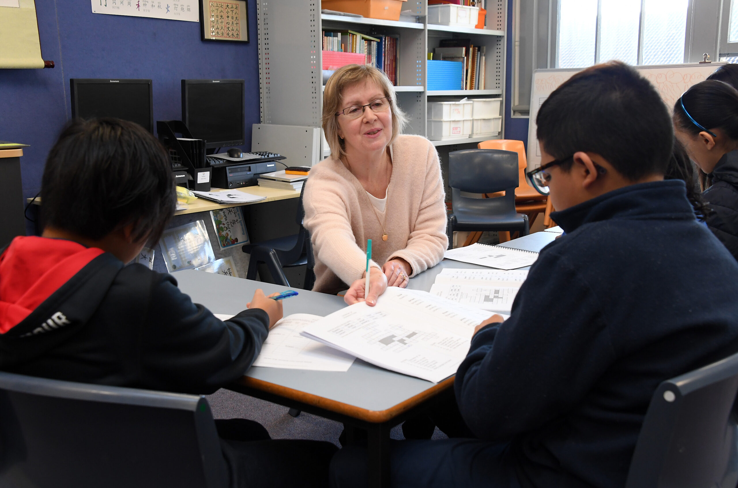 Children sit around a classroom table completing literacy exercises, while their teacher helps.